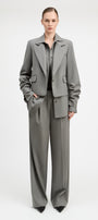 A model stands against a light gray studio background wearing a tailored gray suit with a structured blazer and matching wide-leg trousers, paired with a dark top and black pointed shoes.