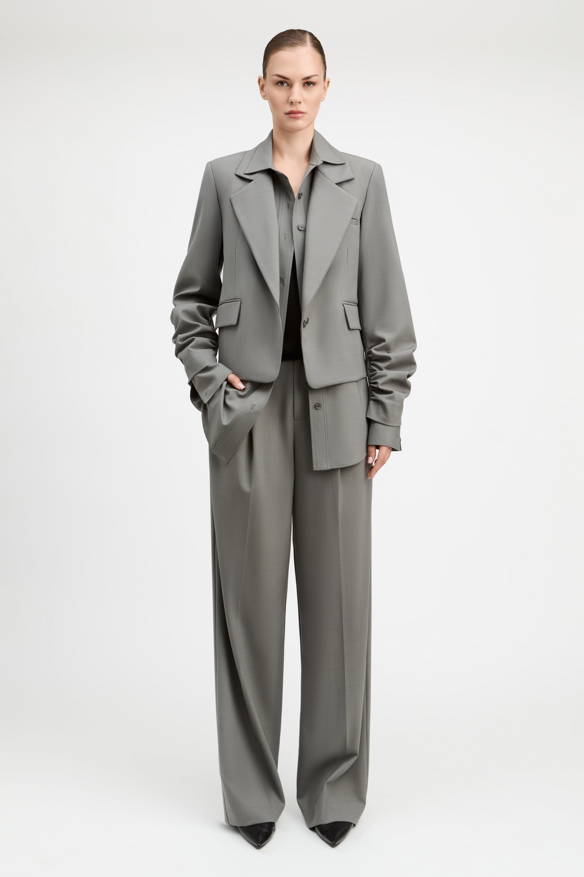 A model stands against a light gray studio background wearing a tailored gray suit with a structured blazer and matching wide-leg trousers, paired with a dark top and black pointed shoes.