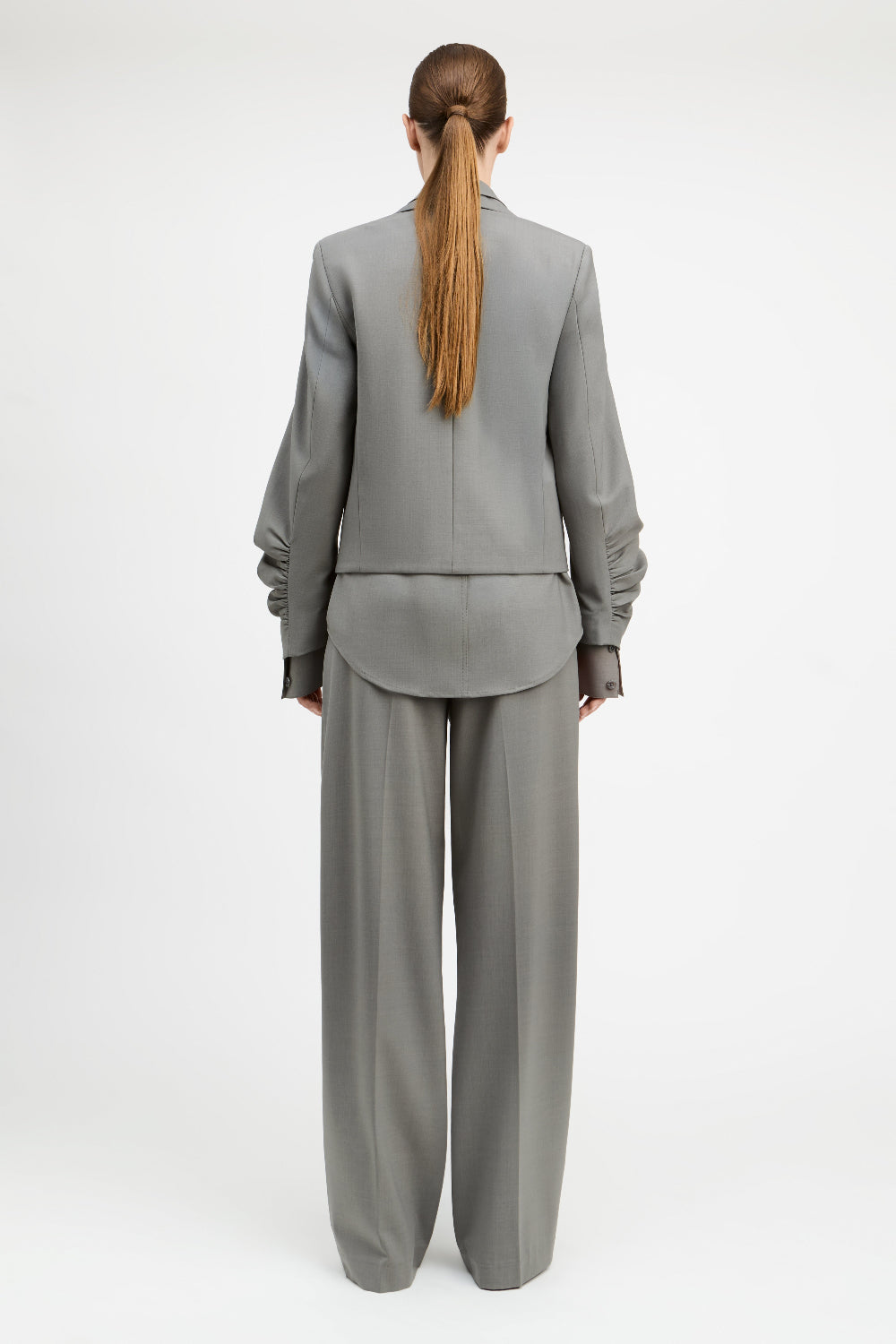 A model viewed from the back wearing a light gray two-piece suit with a structured blazer and wide-leg trousers, set against a plain white studio background.