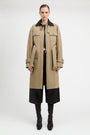 A model stands against a light gray studio background wearing a long beige trench coat with a black collar, black cropped pants, and black knee-high boots.