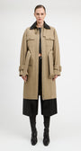A model stands against a light gray studio background wearing a long beige trench coat with a black collar, black cropped pants, and black knee-high boots.