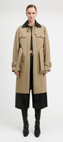 A model stands against a light gray studio background wearing a long beige trench coat with a black collar, black cropped pants, and black knee-high boots.