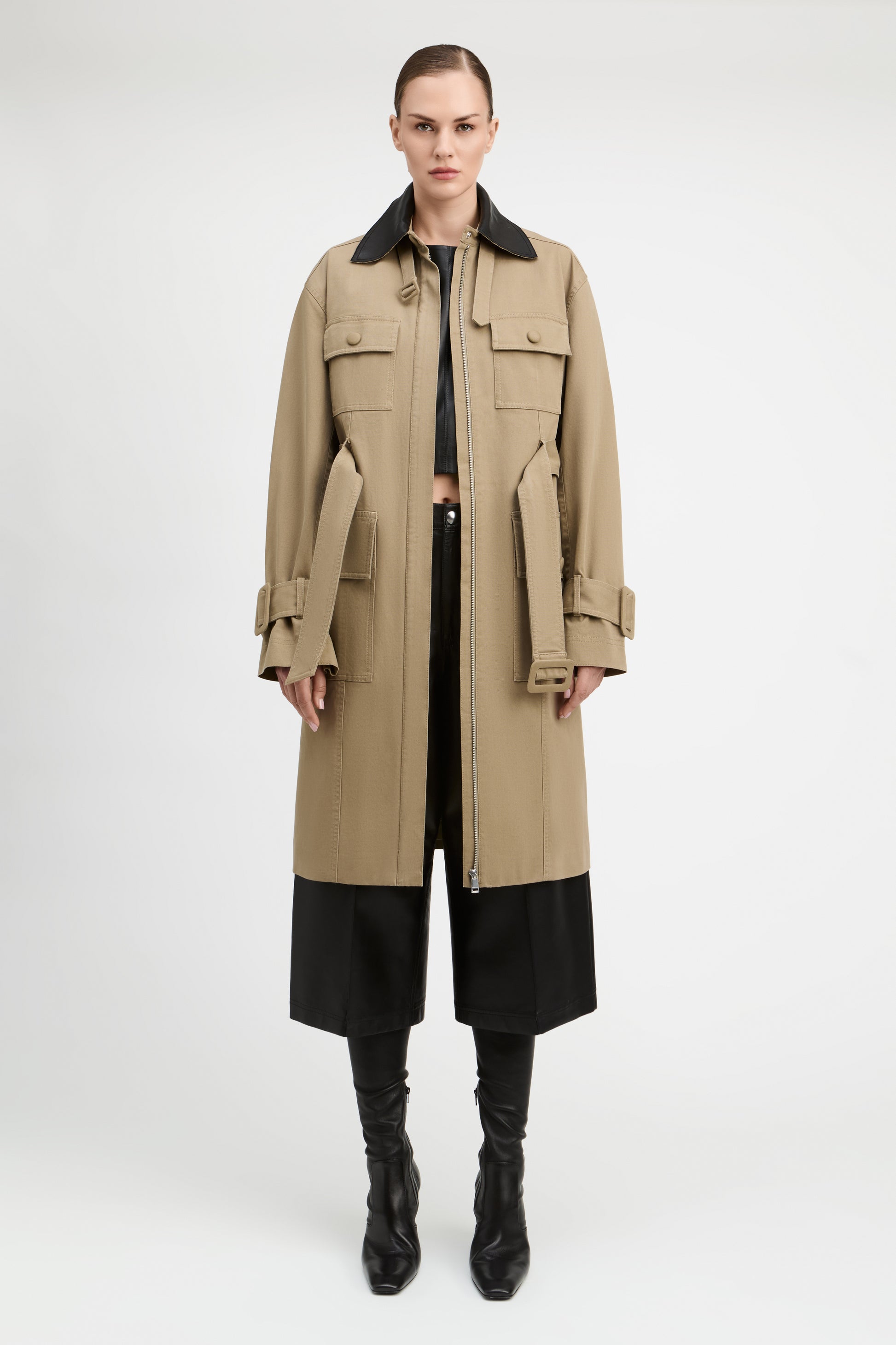 A model stands against a light gray studio background wearing a long beige trench coat with a black collar, black cropped pants, and black knee-high boots.