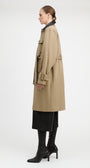A model shown in profile wearing a mid-length beige trench coat over a black skirt and black heeled boots, photographed against a clean white studio background.