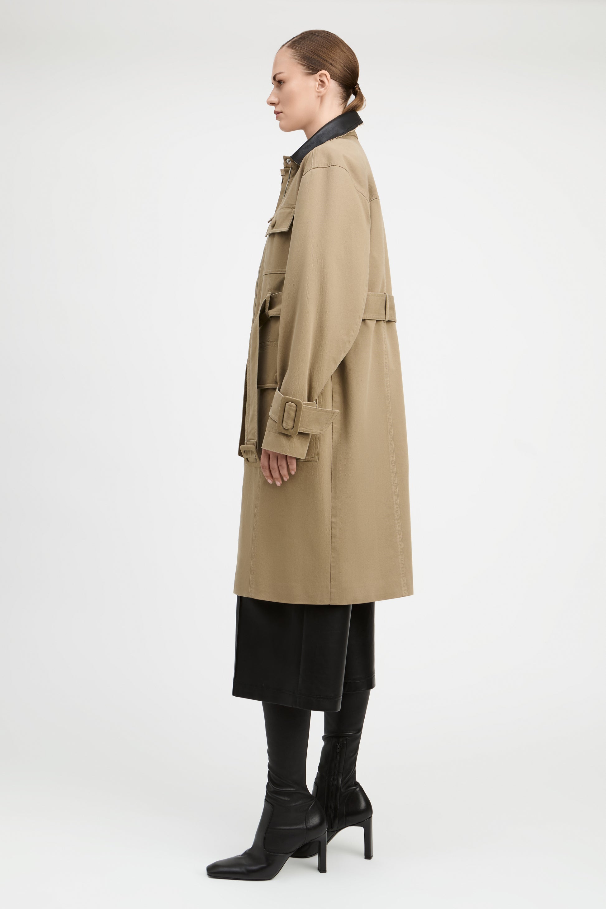 A model shown in profile wearing a mid-length beige trench coat over a black skirt and black heeled boots, photographed against a clean white studio background.