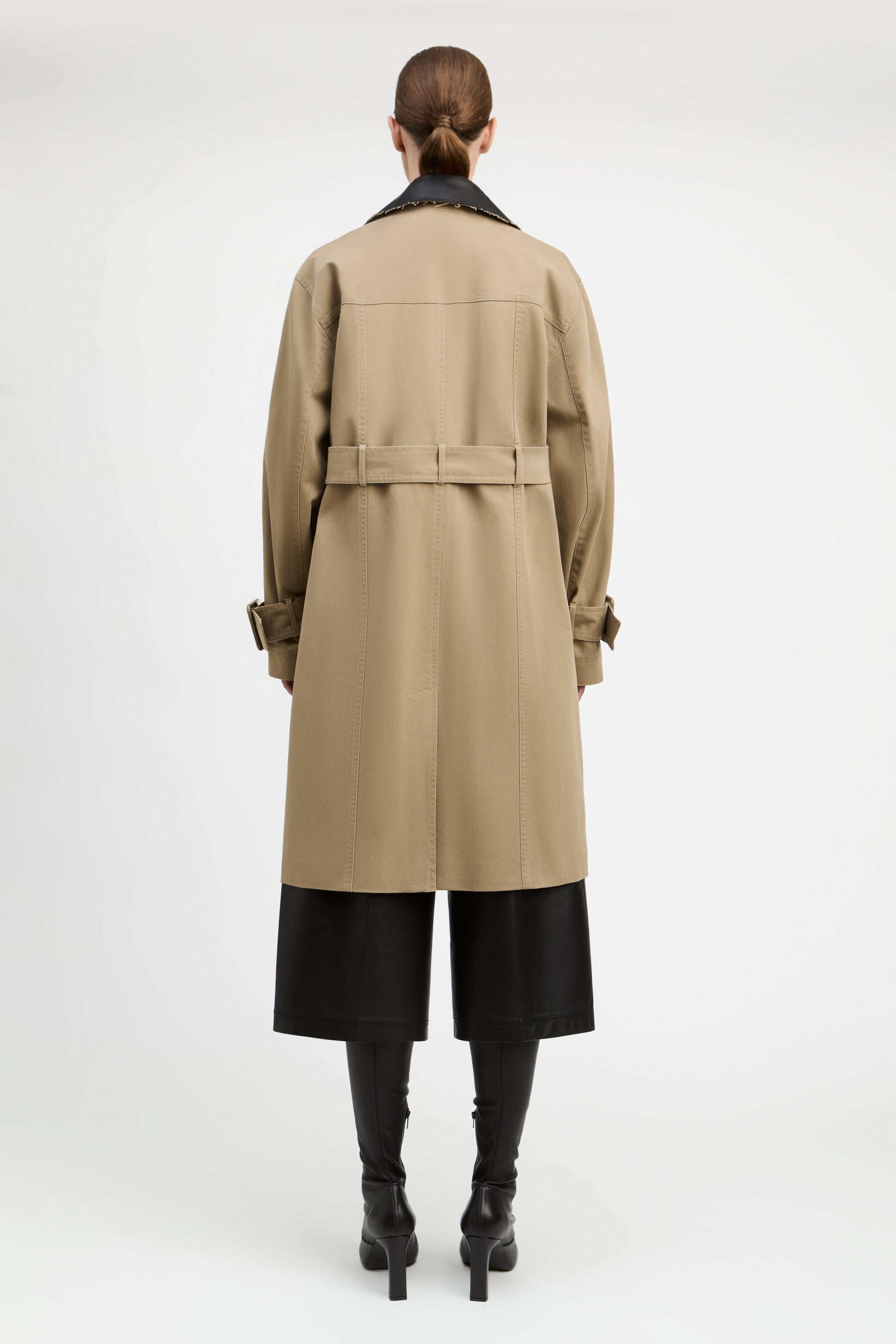 A model shown from the back wearing a mid-length beige trench coat with a black collar, black wide-leg cropped pants, and black heeled boots against a plain light gray studio background.