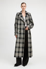A model stands against a light gray studio background wearing a long black and beige plaid trench coat over a white top and black wide-leg pants.
