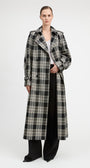 A model stands against a light gray studio background wearing a long black and beige plaid trench coat over a white top and black wide-leg pants.