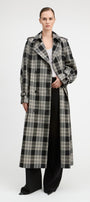 A model stands against a light gray studio background wearing a long black and beige plaid trench coat over a white top and black wide-leg pants.