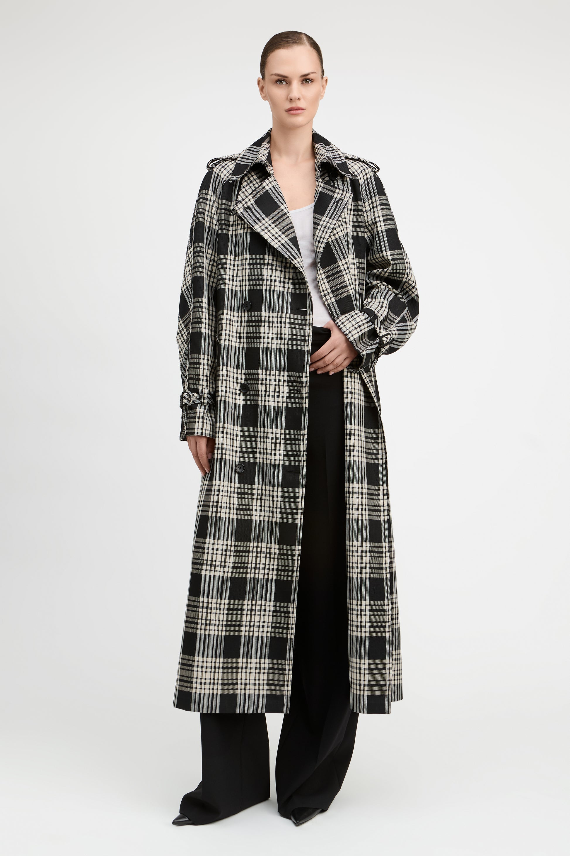 A model stands against a light gray studio background wearing a long black and beige plaid trench coat over a white top and black wide-leg pants.