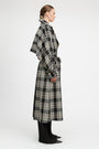 A model shown in profile wearing a long black and beige plaid wrap coat over black wide-leg trousers, photographed against a clean white studio background.
