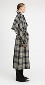A model shown in profile wearing a long black and beige plaid wrap coat over black wide-leg trousers, photographed against a clean white studio background.