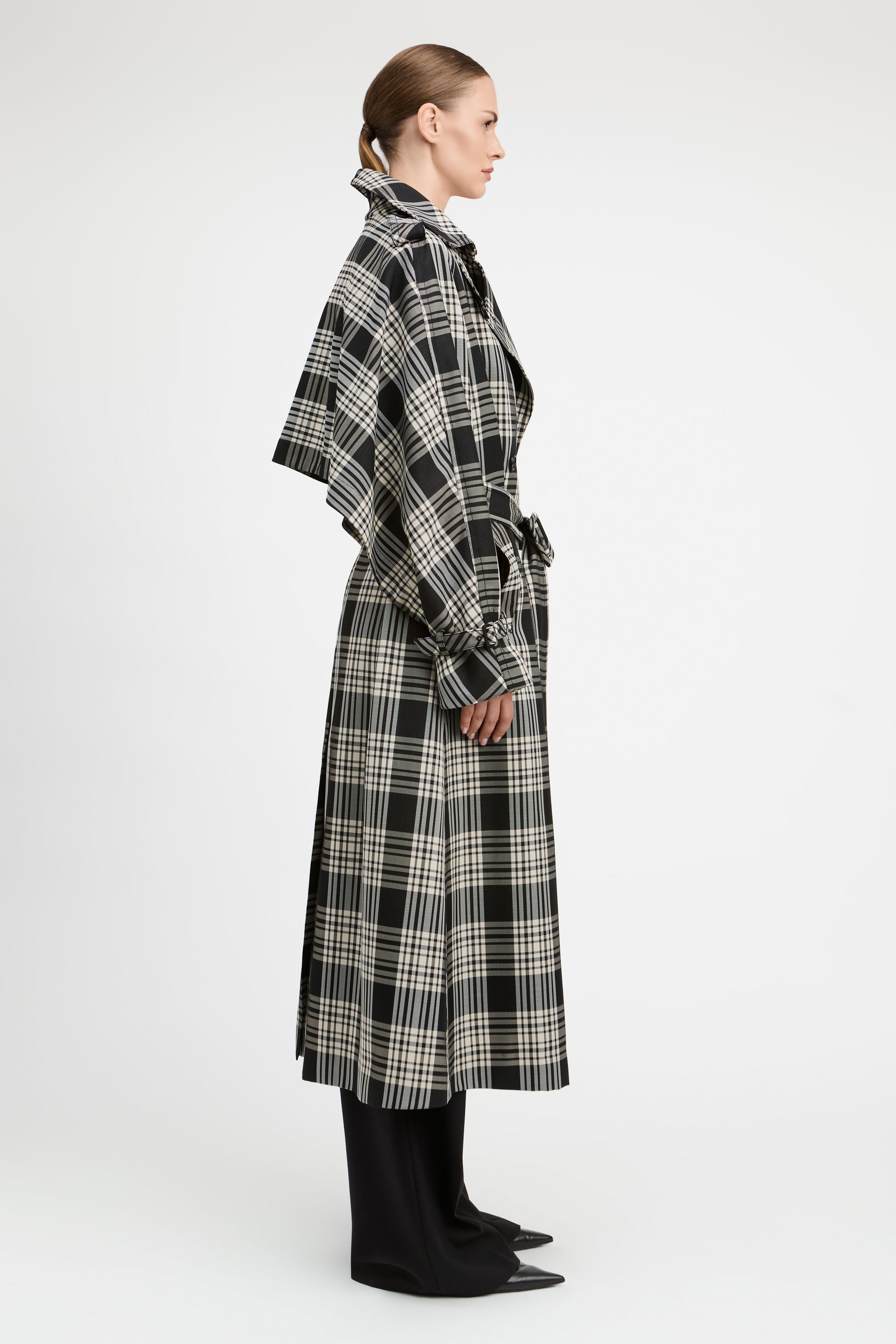 A model shown in profile wearing a long black and beige plaid wrap coat over black wide-leg trousers, photographed against a clean white studio background.