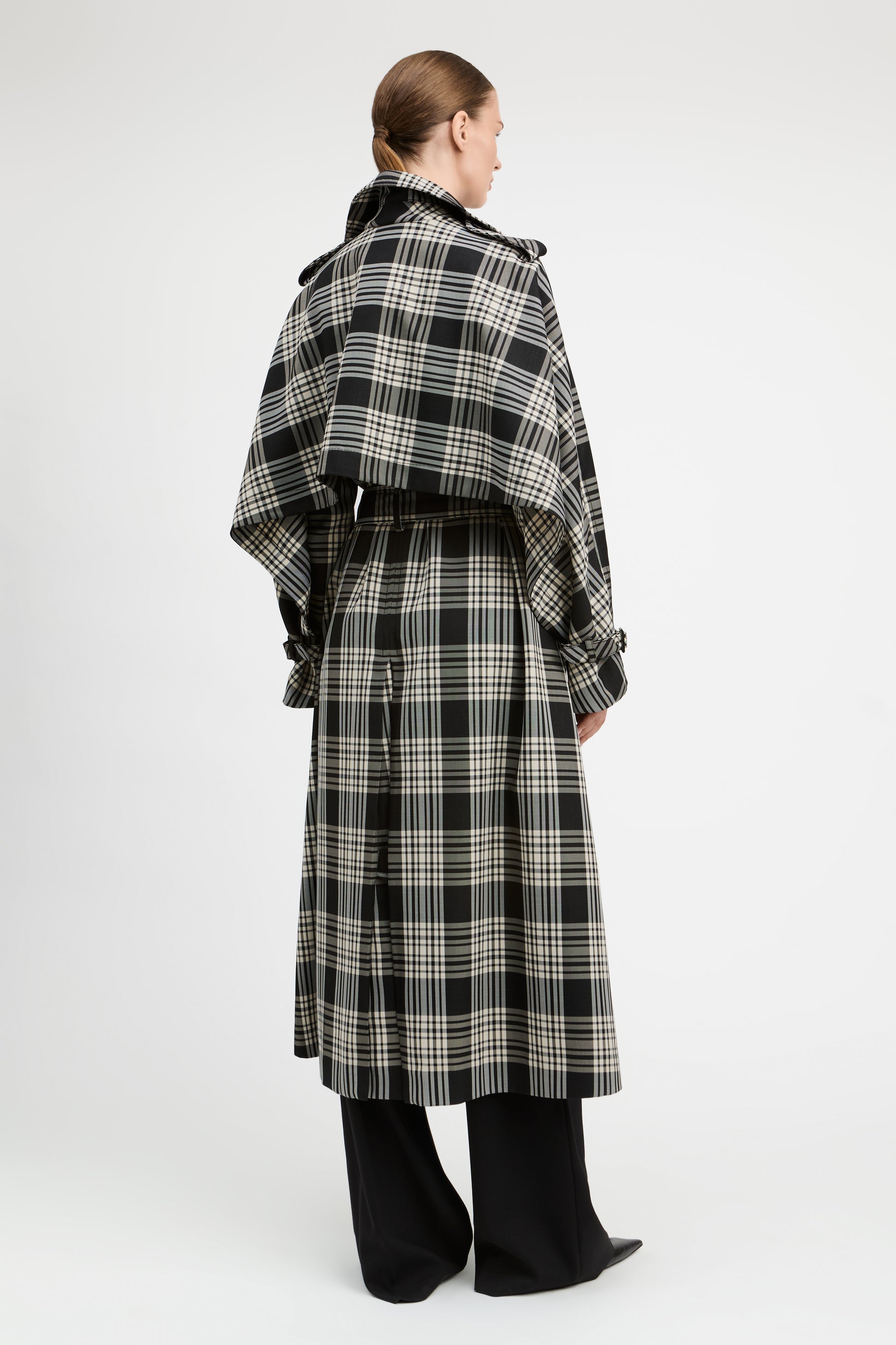 A model shown from the back wearing a long black and beige plaid coat with wide lapels and a self-tie belt, paired with black wide-leg trousers, against a plain light gray studio background.
