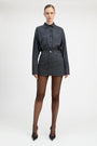 A model stands against a light gray studio background wearing a matching long-sleeve button-front shirt and mini skirt set in a dark blue and black check pattern, paired with sheer black tights and black pointed heels.