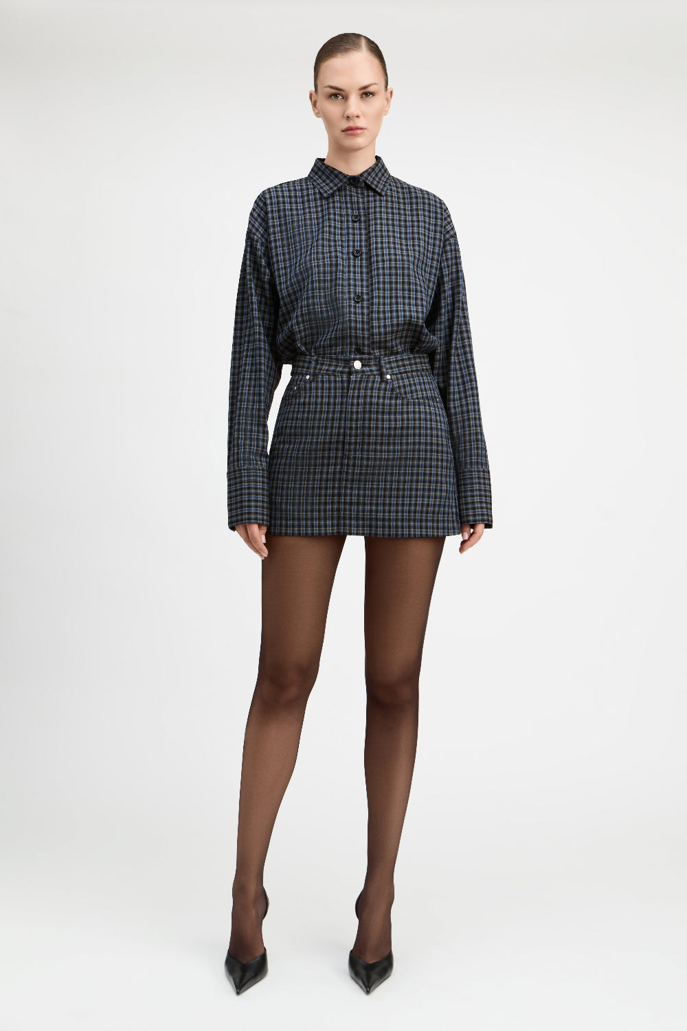 A model stands against a light gray studio background wearing a matching long-sleeve button-front shirt and mini skirt set in a dark blue and black check pattern, paired with sheer black tights and black pointed heels.