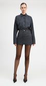 A model stands against a light gray studio background wearing a matching long-sleeve button-front shirt and mini skirt set in a dark blue and black check pattern, paired with sheer black tights and black pointed heels.