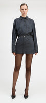 A model stands against a light gray studio background wearing a matching long-sleeve button-front shirt and mini skirt set in a dark blue and black check pattern, paired with sheer black tights and black pointed heels.