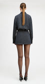 A woman viewed from behind wearing a dark blue and black checkered long-sleeve mini dress with a matching belt, sheer black tights, and black heels, set against a plain white studio background.