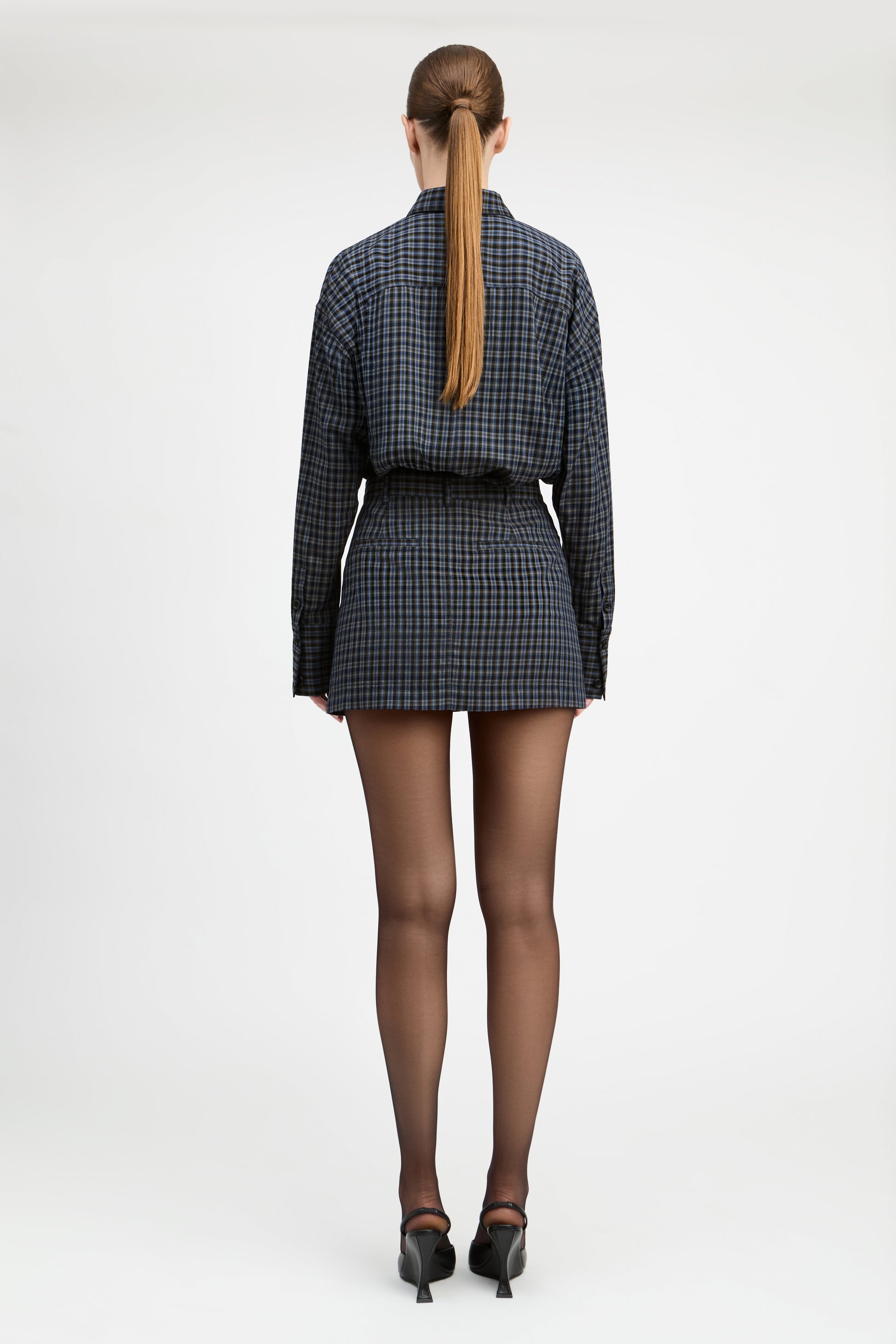 A woman viewed from behind wearing a dark blue and black checkered long-sleeve mini dress with a matching belt, sheer black tights, and black heels, set against a plain white studio background.