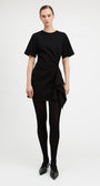 A model wearing a black short-sleeve dress with a twisted front detail and asymmetrical draped hem, paired with black tights and black pointed-toe heels, against a plain light gray studio background.