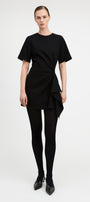 A model wearing a black short-sleeve dress with a twisted front detail and asymmetrical draped hem, paired with black tights and black pointed-toe heels, against a plain light gray studio background.