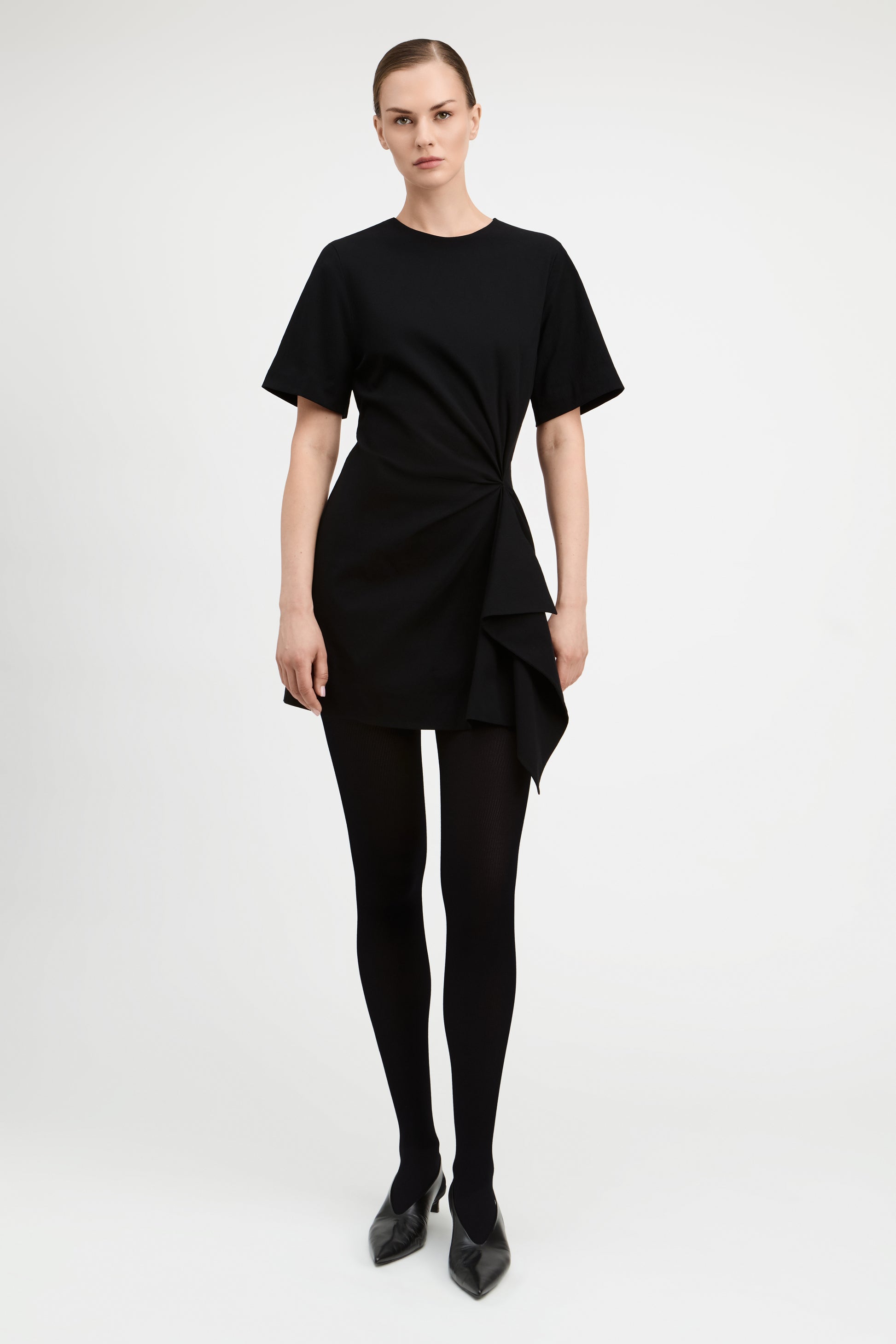 A model wearing a black short-sleeve dress with a twisted front detail and asymmetrical draped hem, paired with black tights and black pointed-toe heels, against a plain light gray studio background.