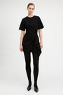 A model wearing a black short-sleeve mini dress with a twisted front detail, paired with black tights and black pointed shoes, photographed against a light gray studio background.