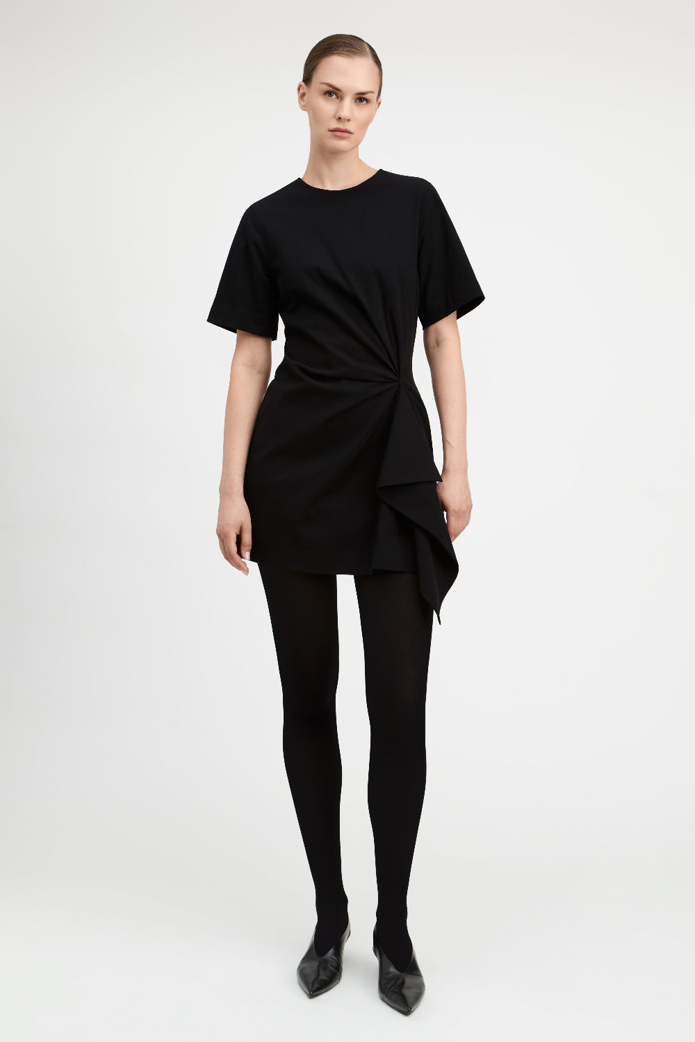 A model wearing a black short-sleeve mini dress with a twisted front detail, paired with black tights and black pointed shoes, photographed against a light gray studio background.