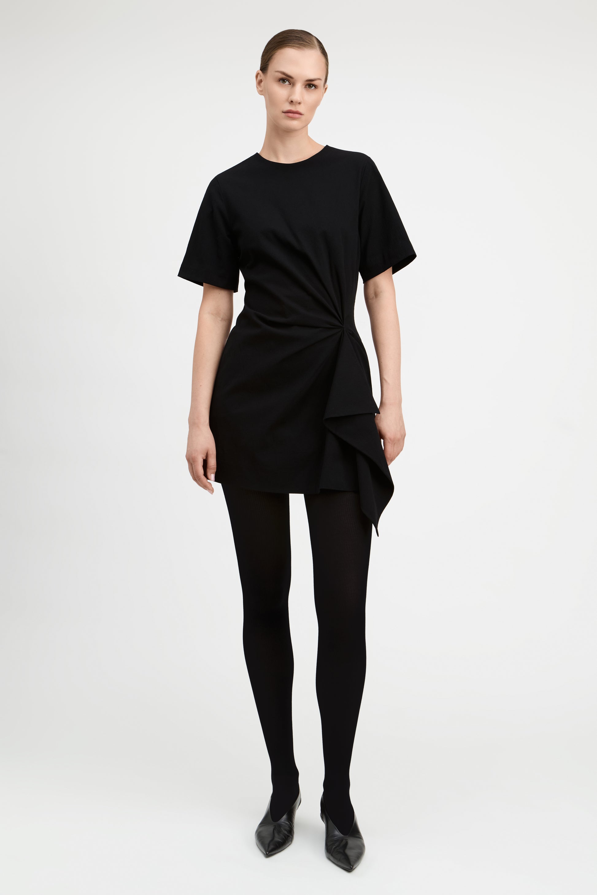 A model wearing a black short-sleeve mini dress with a twisted front detail, paired with black tights and black pointed shoes, photographed against a light gray studio background.