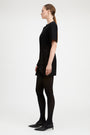 A woman in a black short-sleeve mini dress, black tights, and black pointed-toe heels, shown in profile against a plain light gray studio background.