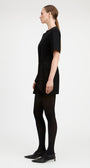 A woman in a black short-sleeve mini dress, black tights, and black pointed-toe heels, shown in profile against a plain light gray studio background.