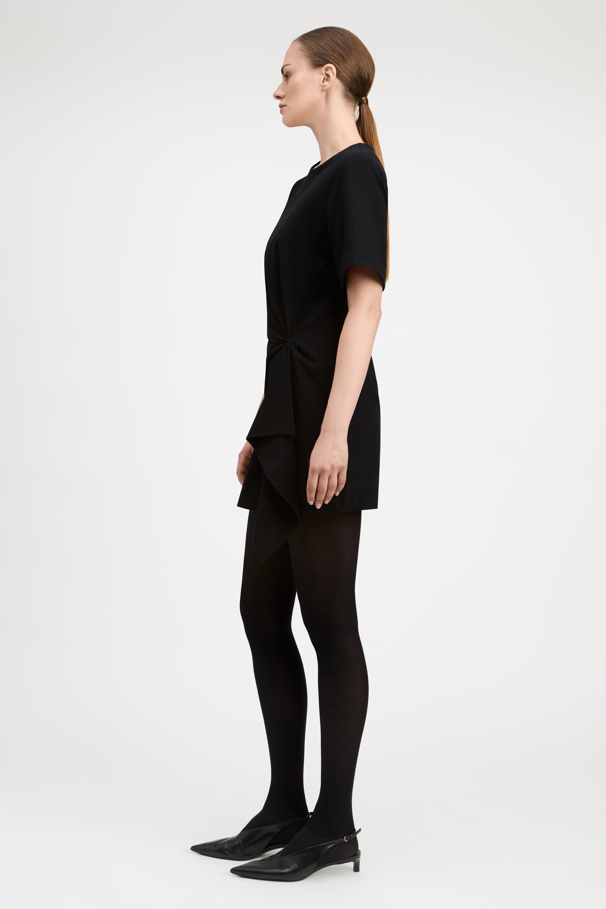 A woman in a black short-sleeve mini dress, black tights, and black pointed-toe heels, shown in profile against a plain light gray studio background.