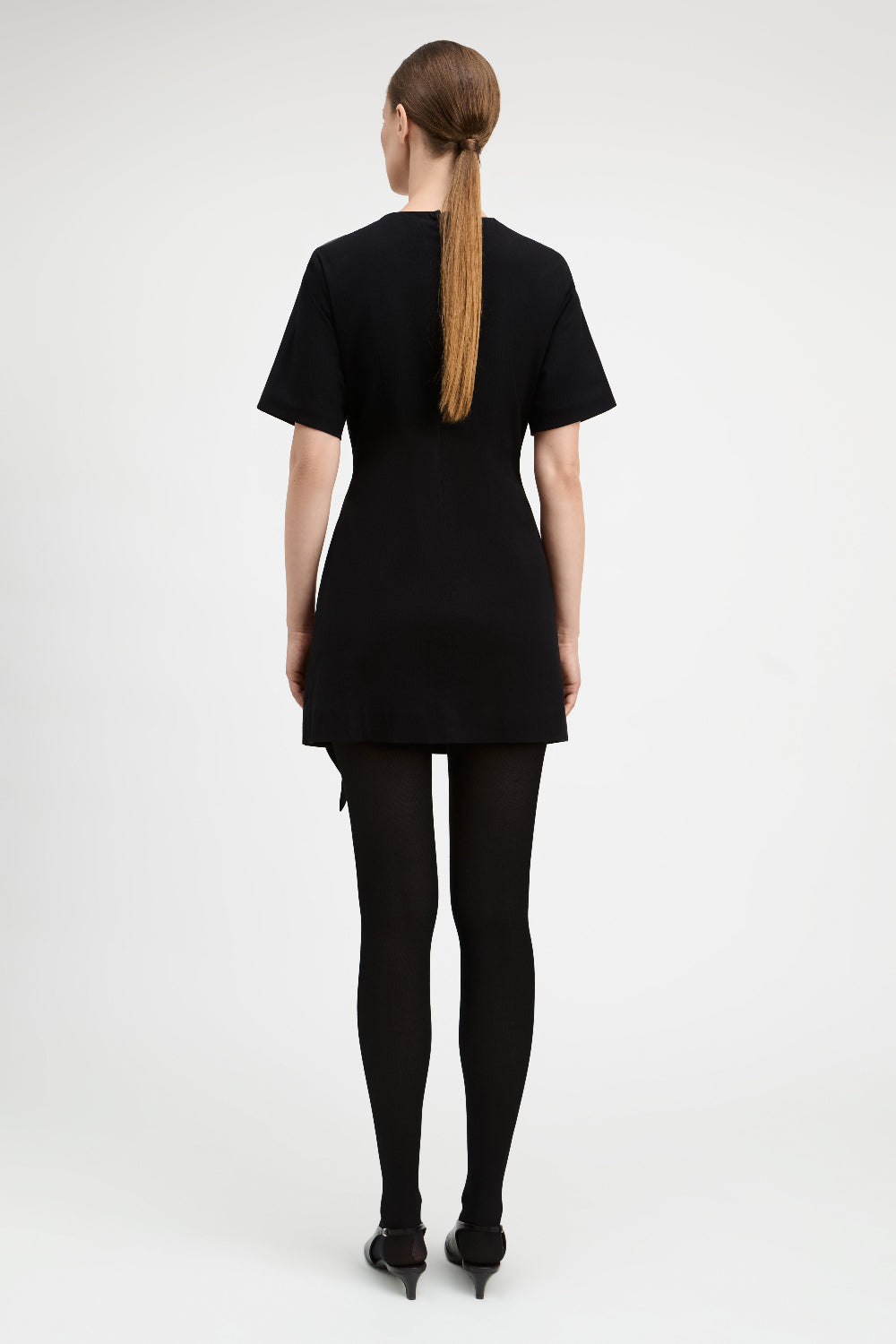 A woman viewed from the back wearing a black short-sleeve mini dress, black tights, and black pointed shoes against a plain light gray studio background.