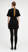 A woman viewed from the back wearing a black short-sleeve mini dress, black tights, and black pointed shoes against a plain light gray studio background.