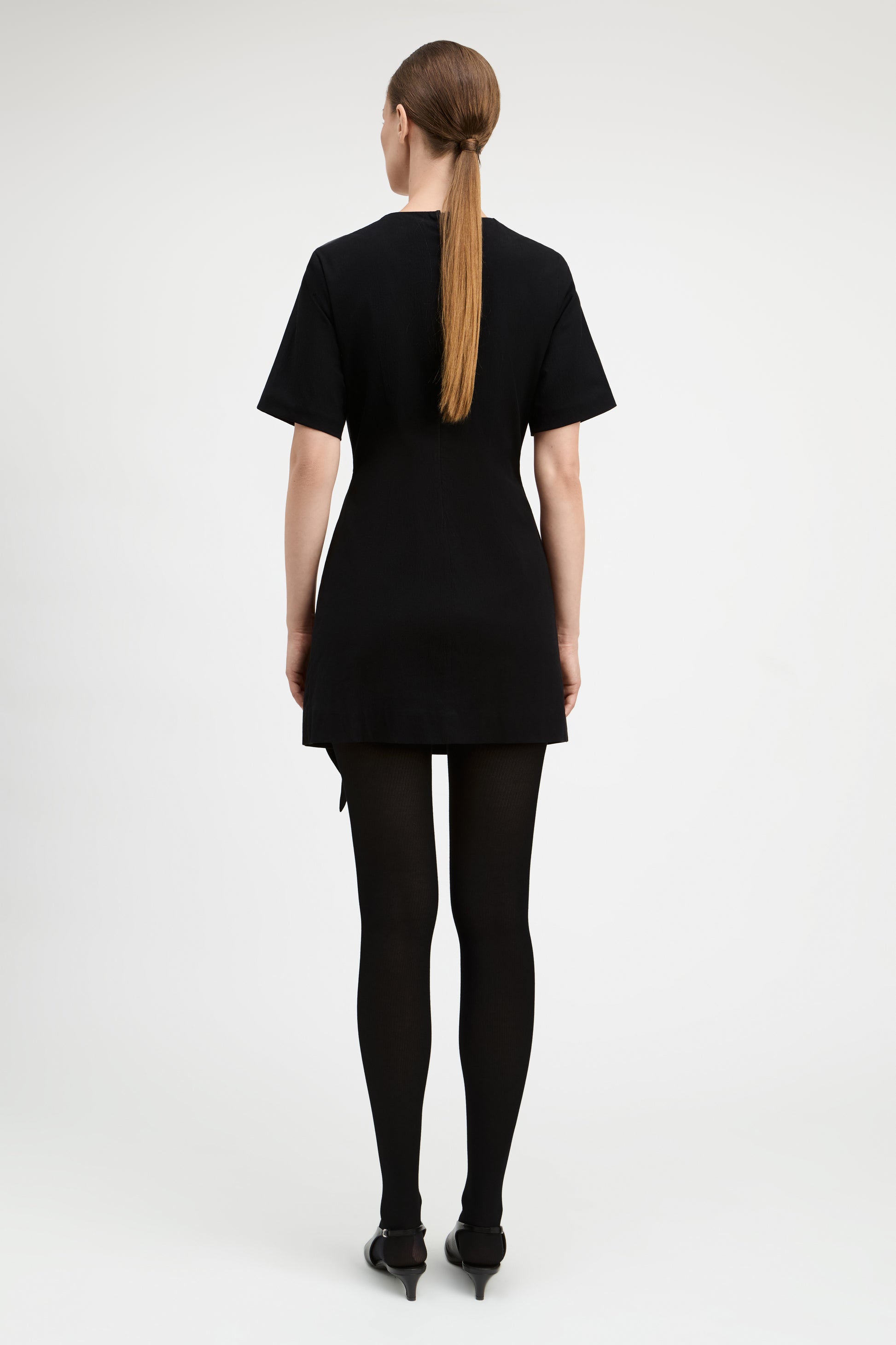 A woman viewed from the back wearing a black short-sleeve mini dress, black tights, and black pointed shoes against a plain light gray studio background.