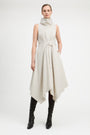 A model wearing a sleeveless, high-neck, asymmetrical midi dress in a light beige or off-white fabric, styled with black knee-high leather boots against a plain white studio background.