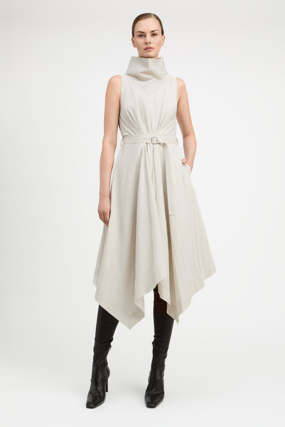 A model wearing a sleeveless, high-neck, asymmetrical midi dress in a light beige or off-white fabric, styled with black knee-high leather boots against a plain white studio background.