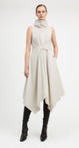 A model wearing a sleeveless, high-neck, asymmetrical midi dress in a light beige or off-white fabric, styled with black knee-high leather boots against a plain white studio background.
