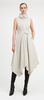A model wearing a sleeveless, high-neck, asymmetrical midi dress in a light beige or off-white fabric, styled with black knee-high leather boots against a plain white studio background.