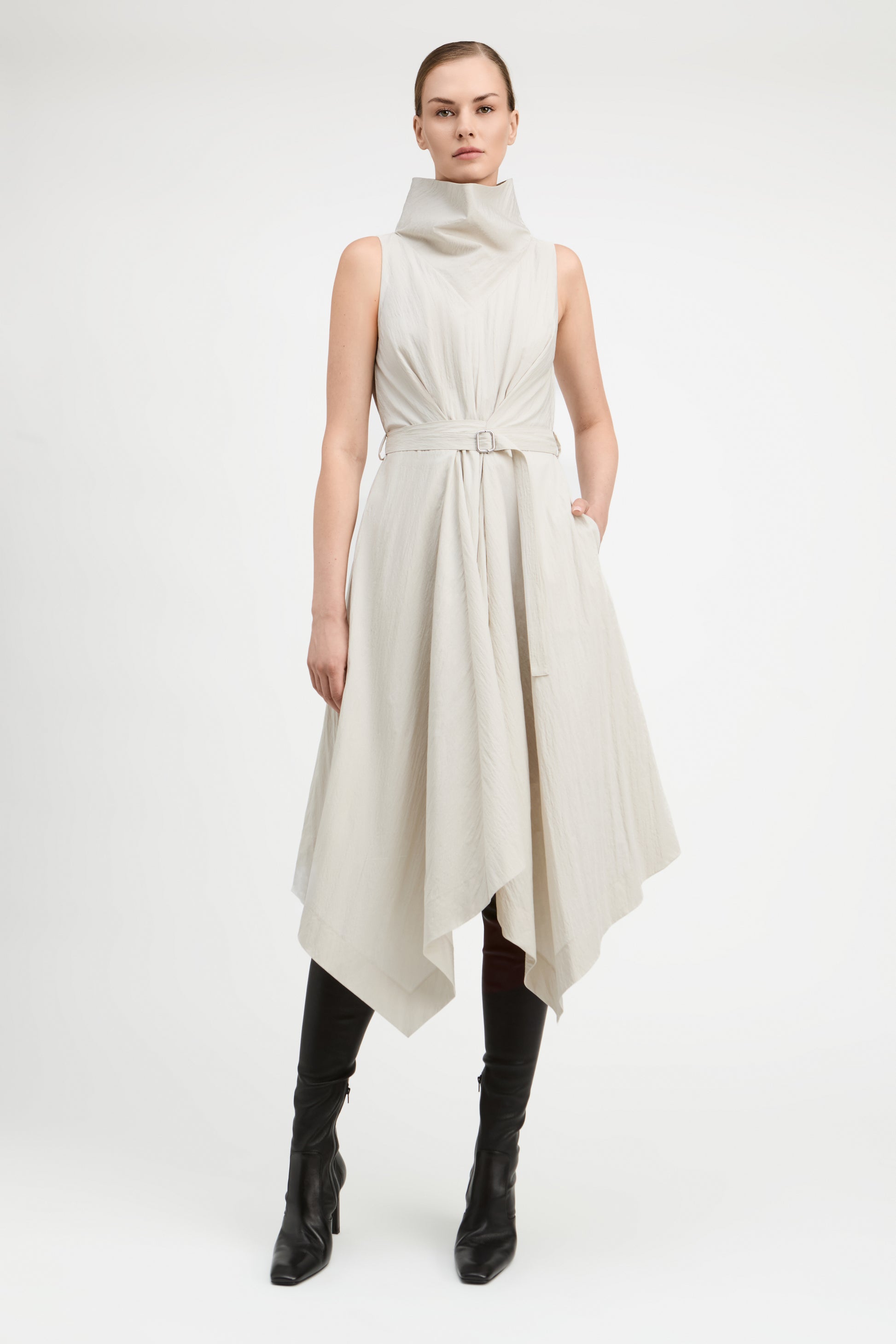 A model wearing a sleeveless, high-neck, asymmetrical midi dress in a light beige or off-white fabric, styled with black knee-high leather boots against a plain white studio background.