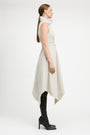 A woman in profile wearing a sleeveless, high-neck, asymmetrical midi dress in a light cream color, styled with black over-the-knee boots against a plain white studio background.