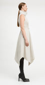 A woman in profile wearing a sleeveless, high-neck, asymmetrical midi dress in a light cream color, styled with black over-the-knee boots against a plain white studio background.