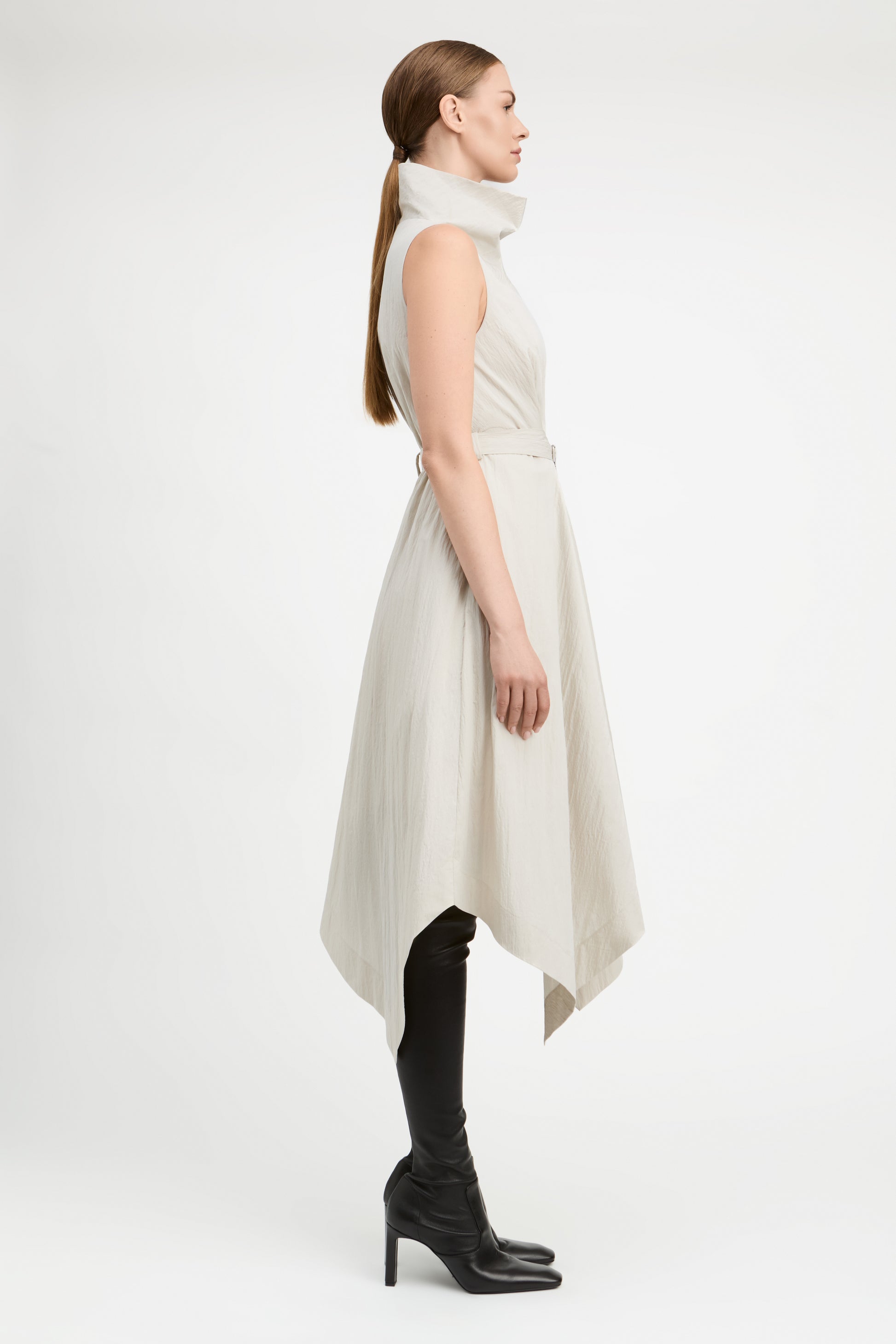 A woman in profile wearing a sleeveless, high-neck, asymmetrical midi dress in a light cream color, styled with black over-the-knee boots against a plain white studio background.