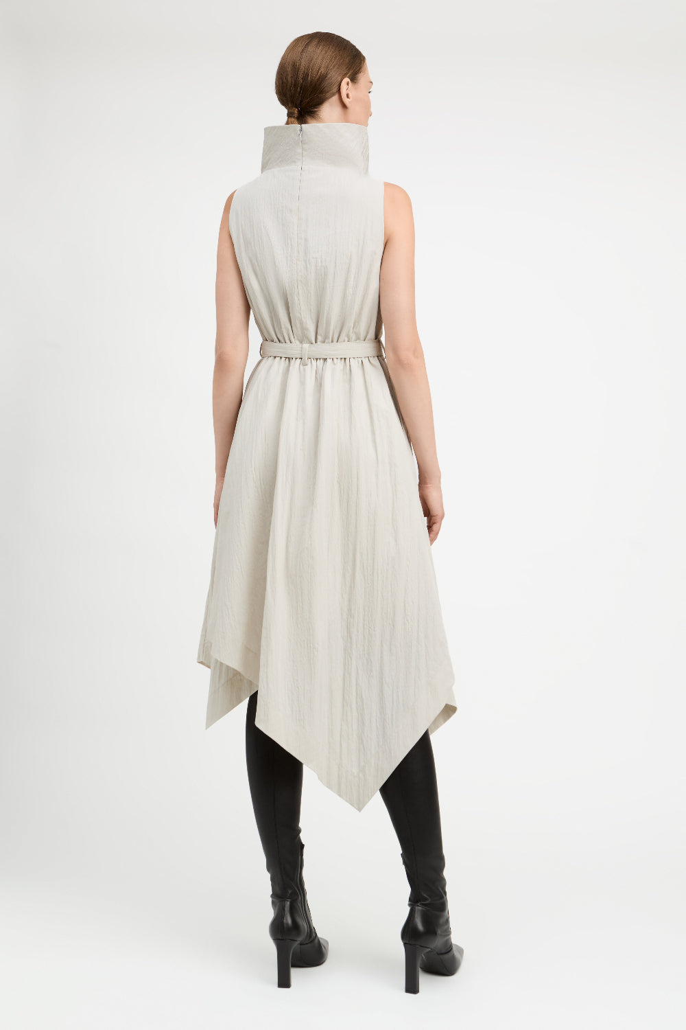 A model viewed from the back wearing a sleeveless, high-neck, belted midi dress in a light beige or off-white crinkled fabric, styled with black knee-high boots against a plain white studio background.