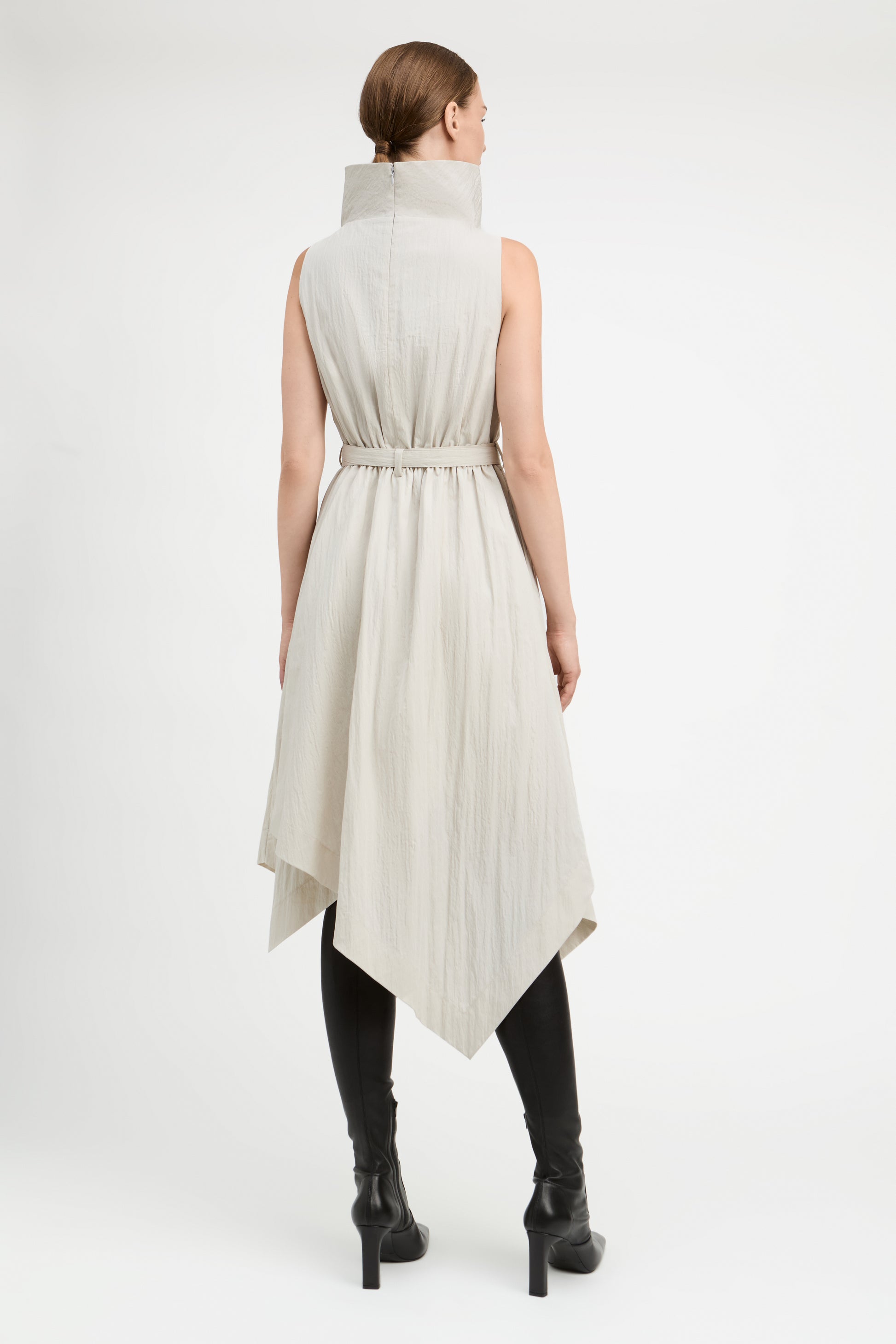 A model viewed from the back wearing a sleeveless, high-neck, belted midi dress in a light beige or off-white crinkled fabric, styled with black knee-high boots against a plain white studio background.