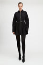 A model wearing a black long-sleeve mini jacket with a front zipper, a matching black belt, black tights, and black pointed-toe heels, photographed against a seamless light gray studio background.