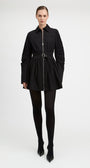 A model wearing a black long-sleeve mini jacket with a front zipper, a matching black belt, black tights, and black pointed-toe heels, photographed against a seamless light gray studio background.