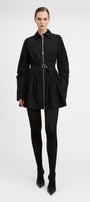 A model wearing a black long-sleeve mini jacket with a front zipper, a matching black belt, black tights, and black pointed-toe heels, photographed against a seamless light gray studio background.