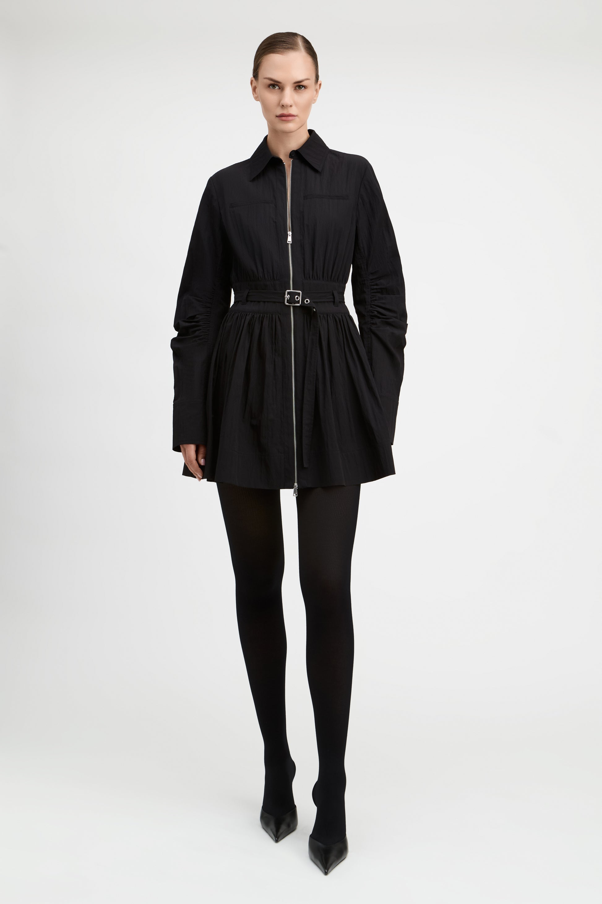A model wearing a black long-sleeve mini jacket with a front zipper, a matching black belt, black tights, and black pointed-toe heels, photographed against a seamless light gray studio background.